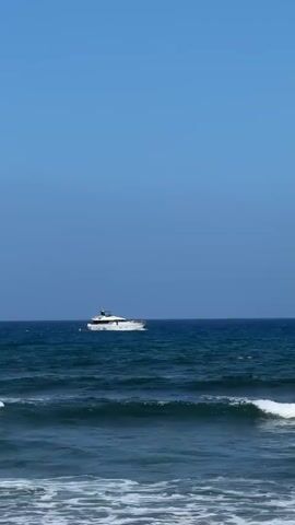 A white yacht with dark trim sails on the choppy blue ocean under a clear blue sky. The camera is positioned low, looking towards the water, with the waves in the foreground. The yacht is moving from left to right across the frame.