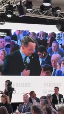 A man speaking into a microphone appears on a video screen. The video is being recorded by someone holding a cell phone. The man is light-skinned with short brown hair and is wearing a dark suit, a light blue and white striped dress shirt, and a blue tie with a repeating pattern. He is wearing a watch on his left wrist and rings on the third and fourth fingers of his left hand. He appears to be speaking at an event called "Ukrainian Breakfast." In the background of the video on the screen are several people seated at tables. Beneath the screen, seated at a table are several more people. A woman to the left of the frame is looking at her phone, and behind her are a video camera and someone working the camera. A man in a blue suit is seated in the middle of the frame. Text on the background indicates that the International Renaissance Foundation is a partner of the event.