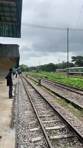 A medium shot pans right across a train station, showing multiple train tracks stretching into the distance. On the left, a covered platform is visible, with a few people standing on it. To the right of the tracks, a long green and white train is stationary. The sky is overcast, and trees and greenery are visible beyond the tracks.