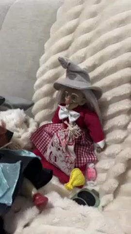 The video shows a doll of an old witch in a red dress with white hair and a gray pointed hat. Another doll of a witch, with long red hair and a blue hat and dress, sits next to her. Both are on a light-colored, fluffy blanket. In the background, a person in pink pajamas is decorating a white Christmas tree with lights and ornaments. A remote control is near the witch doll with the blue hat. The video appears to be taken indoors, in a home setting.