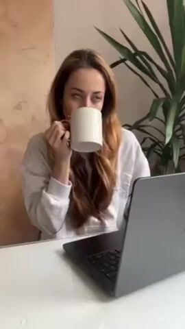 A woman with light brown hair, wearing a light grey sweatshirt, sits at a white desk and drinks from a white mug with a gold handle. A closed grey laptop sits on the desk in front of her. A large potted plant stands to her right. The wall behind her is textured with a light brown and beige pattern. After drinking, the woman smiles briefly at the camera.