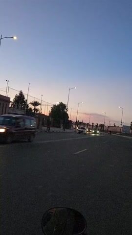 The video captures a twilight street scene from the perspective of a moving vehicle, possibly a motorcycle, indicated by the partial view of a round mirror at the bottom. The road is wet, suggesting recent rain. Several cars with headlights on are visible driving on the road. Along the side of the road are streetlights, trees including palm trees, and a wall or fence. The sky is a gradient of colors, from deep blue to pale pink and orange, indicating dusk. Some figures can be seen walking on the sidewalk.