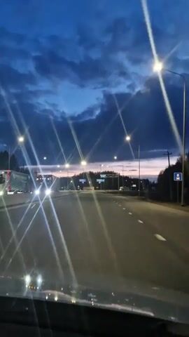The video displays a forward-facing view from a vehicle traveling on a multi-lane highway or major road. The scene is set at dusk or early night, characterized by a deep blue sky with visible clouds. Numerous streetlights illuminate the road, creating prominent starburst effects in the camera's lens. The road ahead appears to be clear with minimal traffic.

Trees and forested areas line both sides of the road. In the distance, several blue road signs with white directional arrows and Cyrillic text are mounted on overhead gantries. Although the text is somewhat obscured by distance and lighting, some signs appear to indicate destinations such as "Вологда" (Vologda) and "Череповец" (Cherepovets), which aligns with the given location context of Selishcha, Russia. The car's dashboard is visible at the bottom of the frame. The overall atmosphere conveyed is calm and peaceful, typical of an evening drive.
