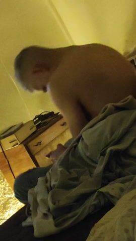 The video shows a man bending over a dresser in a room. The man is shirtless with short, gray hair. He is wearing jeans.  A gray cloth and a portion of a bed are on the right side of the frame. The room is illuminated with a warm light. A keyboard is visible in the upper left corner of the frame. The camera is pointing upwards from a low angle.