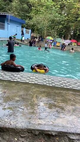 This is a wide shot of an outdoor swimming pool on a cloudy day. The pool water is a vibrant turquoise color. In the foreground, two young children are visible in inflatable tubes. One child, with dark hair, is wearing a black inner tube and appears to be floating. The second child, also with dark hair, is in a yellow inner tube and is looking down. The poolside is tiled with a black and white checkered pattern. Beyond the pool, lush green trees form a dense backdrop. A light blue building with white trim is visible to the left. Several people are scattered throughout the pool, some swimming and others relaxing. There are also tents and umbrellas set up around the pool area, suggesting a recreational or park-like setting. The overall impression is one of a casual, family-friendly atmosphere.