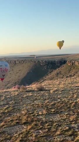 The video captures a serene early morning scene in Yeşilhisar, Turkey, as hot air balloons float over a vast, rugged landscape. The soft golden light of dawn casts long shadows across the undulating terrain, which appears dry and covered in sparse vegetation. In the distance, mountains, some with snow-capped peaks, line the horizon under a clear sky.

Three hot air balloons are visible. The most prominent one in the foreground is white with red and blue accents, clearly displaying the text "Tulip Balloons" along with butterfly and rooster motifs. To its left, another hot air balloon, predominantly yellow with colorful patterns, rises. Further in the background, a third, smaller balloon can be seen. The distinct sound of a hot air balloon burner fills the air, indicating they are either taking off or adjusting altitude. The overall atmosphere is peaceful and majestic, showcasing the beauty of the landscape and the iconic hot air balloon experience.