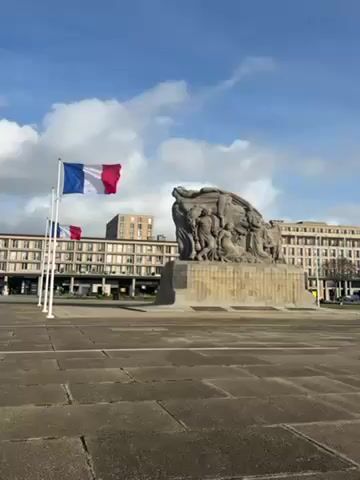 The video pans across a wide, paved plaza with a large, stone monument featuring multiple human figures. Two French flags stand on poles to the left of the monument. Behind the monument, a large building stretches across the background. As the camera pans to the right, it reveals more buildings, a strip of green grass, and a body of water with a modern bridge in the distance. The sky is blue with scattered white clouds. The video appears to have been taken on a bright, sunny day. There are a few people and vehicles visible in the distance, but the plaza itself is largely empty.
