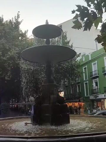 A multi-tiered fountain with water cascading down is the central focus of the video. The fountain is set in a public area with trees and buildings visible in the background. The buildings have a traditional architectural style, with balconies and windows. There are a few people walking around in the background, and some shops with awnings. The overall atmosphere appears to be that of a lively town square.