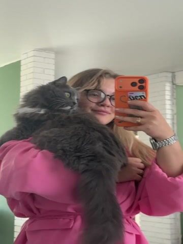 A woman in a pink jacket and glasses is taking a selfie with a fluffy gray cat. She is holding the cat on her shoulder. The background is a room with white brick and green walls. The woman is smiling at her phone, which has an orange case. She is also wearing a silver watch. The cat is looking to the left.