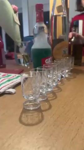 The video shows a bar-style setup, possibly in a home, with a row of small, stemmed glasses lined up on a wooden surface. Behind the glasses are several bottles of different liquids: a bottle of green Get 27 liqueur, a bottle labeled "Création de ...", and another bottle further back. A person wearing a red sweater is visible in the background. A hand comes into frame holding a bottle of clear liquid, with bits of sediment or other material floating within. The hand moves the bottle as if preparing to pour. The overall setting appears casual and relaxed, possibly suggesting a gathering or party.