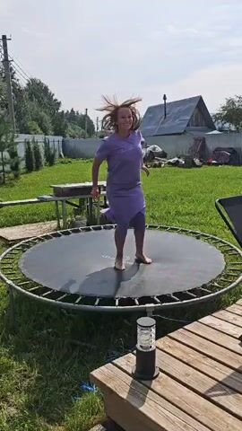 A woman in a purple skirt and t-shirt is captured mid-air as she bounces on a trampoline in a grassy backyard. The trampoline has a black mat and a silver metal frame. Behind her, a wooden fence encloses the yard, and in the background, a house with a dark roof is visible. Sunlight illuminates the scene, casting shadows on the trampoline.
