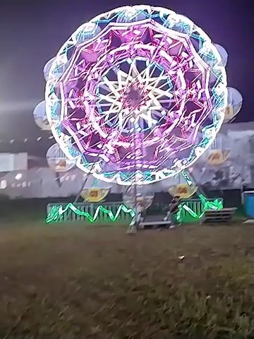 The video shows a night scene at a carnival or fair. The primary focus is a large Ferris wheel adorned with colorful lights. As the camera pans, other attractions come into view, including inflatable slides and various stalls and tents. People are seen walking around, some with children, enjoying the carnival atmosphere. String lights are hung overhead, contributing to the festive ambiance. The ground appears to be grassy.