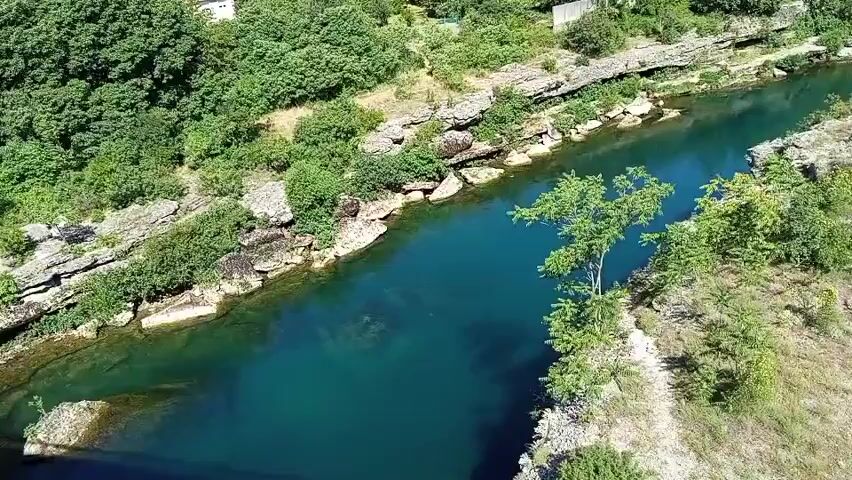 This is an aerial drone shot looking down on a turquoise river flowing between rocky, vegetated banks. The river is wide and appears deep in some areas, with lighter green sections indicating shallower water or visible riverbed. Rocks line the edges of the river, and dense green trees and bushes cover the surrounding hillsides. A single tree with a distinct branch structure grows on the right bank of the river. In the distance, on the upper left side of the frame, a small white building is visible amidst the foliage. The overall scene depicts a natural, scenic landscape with a vibrant river.