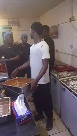 The video shows a group of people in what appears to be a commercial kitchen. One person is holding a large metal pan filled with a reddish-brown substance, possibly food. Several other individuals are visible in the background, some wearing hairnets, suggesting they are working in a food service environment. The kitchen appears to be functional, with various appliances and equipment present, including refrigerators and a stove top. The lighting is somewhat dim, giving the scene a utilitarian feel. The focus of the video is on the person with the pan, who seems to be presenting or moving it.
