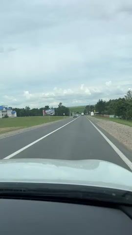 The video shows a first-person perspective of driving on a paved road. The road has two lanes in the direction of travel, separated by a solid white line, and a dashed white line on the left. The weather appears overcast with some clouds. To the left of the road, there is a grassy field with some trees and buildings in the distance. A billboard is visible on the left side of the road. To the right of the road, there are trees and a gravel shoulder with a red and white barrier. Several cars are visible on the road, some in the distance and one passing by. The dashboard of the vehicle is partially visible in the foreground. The scene appears to be a rural or semi-rural setting.