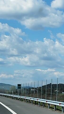 This media file captures a view from a moving vehicle on a highway, likely in Romania. The scene is dominated by a bright blue sky filled with fluffy white clouds, indicating fair weather and possibly a sunny day, though the light suggests it could be morning or late afternoon. The highway itself is paved and has clear white lane markings. A metal guardrail runs along the side of the road, separating it from a sloping embankment covered in vegetation.

In the distance, rolling hills or mountains are visible, partially obscured by the haze of the day. There are also some signs visible along the highway. One green sign shows destinations and distances, including "Sibiu," "Curtea," and "Arges." Another smaller black sign is also present, but the text is too blurry to be legible. The overall impression is one of travel and movement through a scenic, rural landscape. There are no people visible in the frame. The camera perspective is looking forward and slightly to the right, giving a sense of the road ahead.