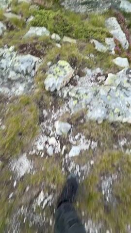 The video shows a person's feet walking on a rocky and grassy mountain trail. The camera pans up to reveal a vast mountain landscape with rolling hills covered in green vegetation, scattered rocks, and a small lake in the distance. The sky is a pale blue with a few scattered clouds, and the light suggests it might be early morning or late evening. There are small pink flowers blooming on the mountainside. The overall scene is one of natural beauty and rugged terrain.