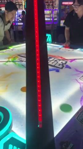 Four people are playing air hockey at an arcade. Two men are prominently featured, actively engaged in the game on a brightly lit table with colorful graphics. One man, wearing glasses and a dark shirt, is intensely focused, leaning over the table. The other man, in a light grey shirt, is also playing and is seen smiling briefly. A third player, whose back is to the camera, is on the opposite side of the table. The air hockey table has "OPEN ICE QUAD-AIR" printed on its surface. Scoreboards above the table display player scores, with one score showing "PLAYER 1" at "15" and "PLAYER 2" at "11" earlier. Other arcade machines are visible in the background.