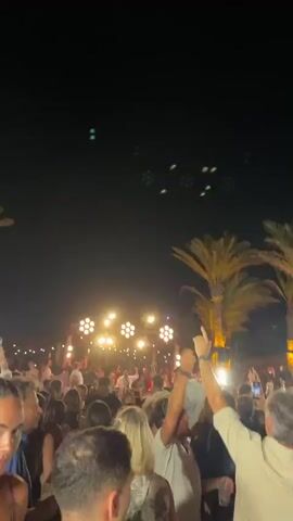 The video shows a crowded outdoor event at night. Palm trees are visible on the right side of the frame, illuminated by lights. In the background, there is a structure with lights and many people gathered. The foreground is filled with the backs of people, some with their arms raised, suggesting they are dancing or celebrating. The overall atmosphere appears to be a lively party or festival.
