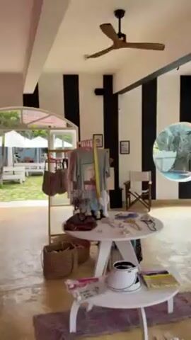 The video depicts an interior view of a retail store, displaying merchandise on round tables and racks. The store has a modern aesthetic with black and white striped walls, a light-colored concrete floor, and a ceiling fan. An open doorway leads to an outdoor patio with white picnic tables, umbrellas, and green grass. A person walks across the patio, visible through the doorway. The store features items such as t-shirts, bags, jewelry, and other accessories.