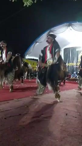 The video shows a group of men performing a traditional dance outdoors at night. They are dressed in elaborate costumes featuring black tops, patterned leg coverings, and white headbands. Several dancers are holding wooden sticks and appear to be performing synchronized movements. In the background, a white tent is visible, and there are lights illuminating the performance area. Some of the dancers have facial hair, specifically mustaches. The ground is covered with a red carpet and dirt. The overall atmosphere suggests a cultural performance or festival.