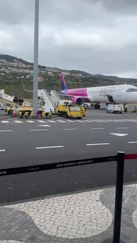 The video showcases an airport setting, specifically the tarmac area with planes, ground vehicles, and airport infrastructure. The initial shot presents a Wizz Air plane, painted white with pink and purple accents. Adjacent to it is a yellow tow vehicle and a set of stairs leading to the plane's entrance. In the background, mountainous terrain covered with residential buildings is visible. The camera then shifts its focus to capture a Ryanair plane, characterized by its blue and yellow livery. People, presumably airport staff, are observed working around the planes. The view transitions again, this time showing the scene through the airport terminal's architecture. The camera pans from behind a white pillar, revealing more of the tarmac, airport vehicles moving in the distance, and additional pillars lining the terminal. The weather appears overcast.