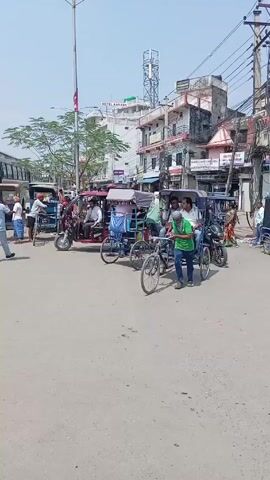 The video shows a busy street scene in what appears to be a town square in Asia. Several auto rickshaws, some with passengers, are clustered near the center of the frame. A man in a green shirt is pushing a three-wheeled cycle rickshaw, carrying two passengers. Other people walk around, and motorcycles and a large bus are also visible. Buildings line the street, with signs in a language other than English. The sky is light blue and the weather appears sunny. The scene is dynamic with people and vehicles moving around.