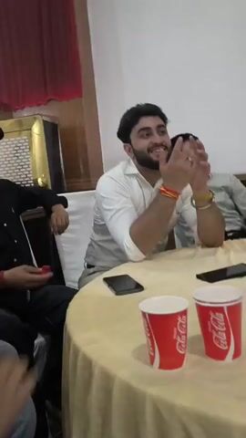 The video begins with a medium shot of a man sitting at a round table covered with a beige tablecloth. He is wearing a white collared shirt and has dark hair and a beard. He is smiling and clapping his hands. Two red cups with Coca-Cola branding are visible on the table in front of him, along with two smartphones. Behind him, another man in a light-colored shirt is visible, also smiling. The environment appears to be an indoor event space with white chairs covered in white fabric and draped with white cloth. Red curtains are visible in the background, as is a television screen.

The video then transitions to a close-up of a person holding a smartphone. The background is blurred, but it appears to be another indoor event setting with people and covered tables.

The video then cuts to a medium shot of a man with curly dark hair, wearing a black jacket over a white t-shirt and khaki pants. He is looking down at a smartphone he is holding. He appears to be in a room with white chairs, some of which are covered with white fabric. Balloons are visible in the background.

The video then shows a low-angle shot of rows of chairs covered in white fabric. Some chairs are unoccupied, while others have people seated in them, mostly seen from the back. Tables covered in light-colored tablecloths are visible between the rows of chairs. The floor is tiled.

Finally, the video shows a blurry, handheld shot of people walking through a dimly lit room. The people are mostly visible from the back or side. Some appear to be women, wearing dresses or traditional Indian attire. The environment seems to be a larger event space with tables and chairs.
