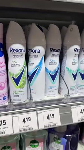 The video shows a close-up shot of a shelf in a store displaying various deodorants. The deodorants are arranged in rows, with some products closer to the camera than others. The brand names visible include Rexona, Nivea, Sanex, and Fa. The products are in spray cans of various colors and designs. Some cans have price tags in front of them. The shelf is well-lit and appears to be part of a retail environment. The focus is on the product display, with no people visible in the frame. The video is shot from a slightly elevated angle, looking down at the shelf.