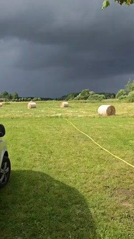 A light-colored van is parked on the left side of the frame in a grassy field. Several round hay bales are scattered across the field. The sky is dark with heavy clouds, indicating an approaching storm. A yellow hose runs across the grass from the van towards the hay bales.