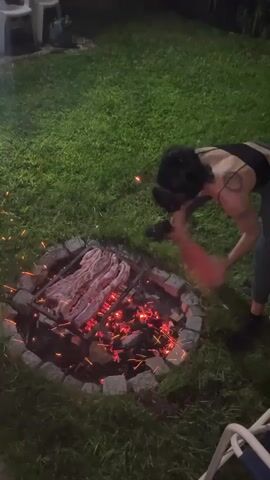 A woman is tending a backyard grill. A fire pit made of gray stones holds a metal grill over red coals, and several pieces of meat lie on the grill. The woman is wearing a black top, gray pants, and black shoes. Her hair is pulled back in a bun. She is using a flat, cardboard piece to fan the fire. Sparks fly upward. The grill is set on a lawn of green grass. A white plastic chair is on the left and a blue and white chair on the right. It is dusk or nighttime.