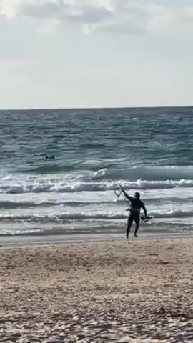 A person in a dark wetsuit walks into the ocean at a beach. They carry a board in their left hand and hold a tall thin piece of equipment above their head in their right hand. The ocean has small waves coming to shore. At least two other people are further out in the water, either swimming or using watercraft. The sky is mostly clear with a few wispy clouds. The beach is sand colored.