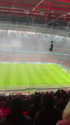 The video shows a soccer match in a large stadium, with many spectators in the stands. The field is green, and players in white and red jerseys are visible. There is fog or smoke obscuring parts of the view. The camera appears to be held by a spectator, and the audio captures the sounds of the crowd.
