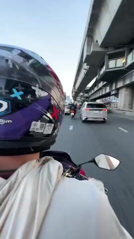 A first-person perspective video shows a person riding a motorcycle in traffic. The rider is wearing a helmet with a purple and black design featuring crosses and cartoon-like figures. The rider's hands are visible on the handlebars, wearing white fingerless gloves and a light-colored jacket. The motorcycle's digital display shows the speed increasing from 28 to 35. The environment is an urban setting with an elevated train track or highway structure overhead. Several cars and other motorcycles are in front of the rider, moving in the same direction. The sky is clear and appears to be late afternoon or early evening. The road is asphalt with white lane markings.