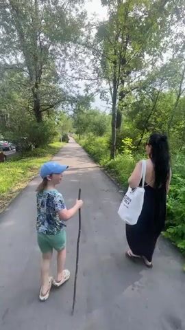 Two people walk along a paved path lined with trees and green foliage. The person in front, seen from behind, is a child wearing a blue baseball cap, a patterned t-shirt, and green shorts, and is holding a long stick. The person behind is an adult wearing a black dress and carrying a white tote bag. The adult is looking to the right, away from the child. The path appears to be in a park or natural setting.