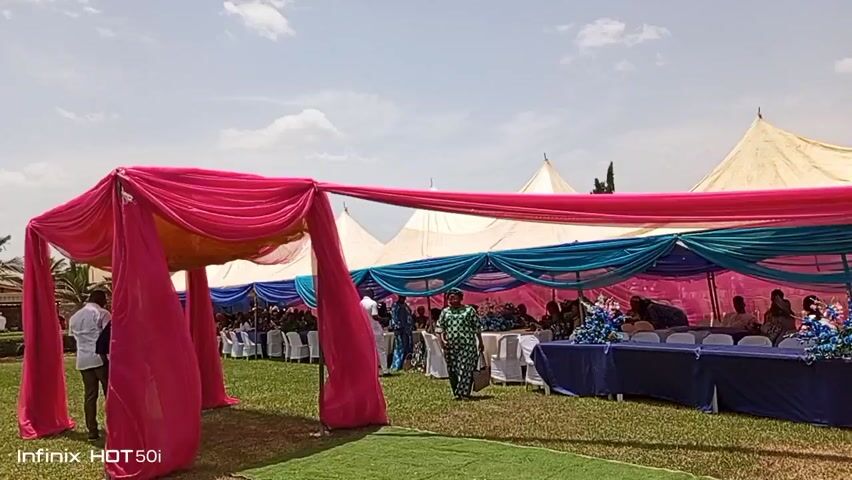 The video captures an outdoor event, likely a wedding or celebration, set on a grassy lawn under a partly cloudy sky. A vibrant pink tent-like structure stands prominently in the foreground. Beyond this, a larger tent is adorned with layers of pink, teal, and white draping. Tables are set up beneath the tent, with people seated, suggesting the event is in full swing. Several individuals are walking around the lawn, including one in a patterned green dress carrying a bag. Palm trees and a building can be seen in the background, adding to the outdoor setting. The video’s perspective is from a stationary position, allowing for a clear view of the event setup and attendees. The camera’s lens flares slightly, indicating strong sunlight. An "Infinix HOT 50i" watermark appears in the lower left corner.