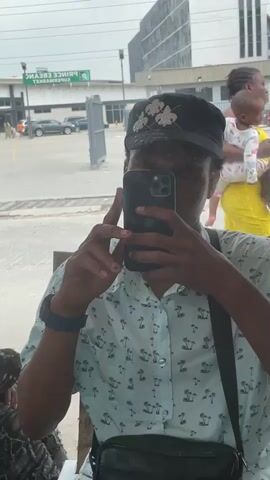 A person takes a selfie in a mirror. The person is wearing a light blue patterned shirt and a black cap with floral embellishments. The person is holding a smartphone and making hand gestures. In the reflection, a street scene with cars and buildings is visible, including a sign that reads "PRINCE EBANO SUPERMARKET".
