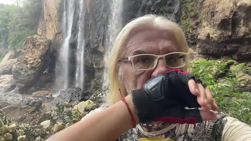 An older man with long blond hair and a white beard is standing in front of a waterfall. He is wearing glasses, a printed shirt, and a glove on his left hand. The background is lush with vegetation and rocks. The man speaks.