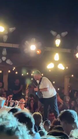 A man in a white t-shirt and dark pants stands on a stage, holding a guitar and singing into a microphone. He is surrounded by a crowd of people, many of whom are holding up their phones to record. The stage is lit by a series of bright spotlights and string lights. In the background, there is a dark sky and what appears to be a building or structure. The overall atmosphere is one of a live musical performance or concert.
