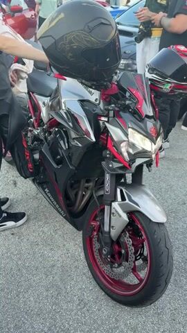 The video shows a close-up view of a black and red Kawasaki motorcycle parked on asphalt. A black helmet with a gold design sits on the motorcycle's seat. The motorcycle's headlights are on. In the background, several people are visible, some holding phones, and other vehicles are parked. The scene appears to be an outdoor gathering or event.