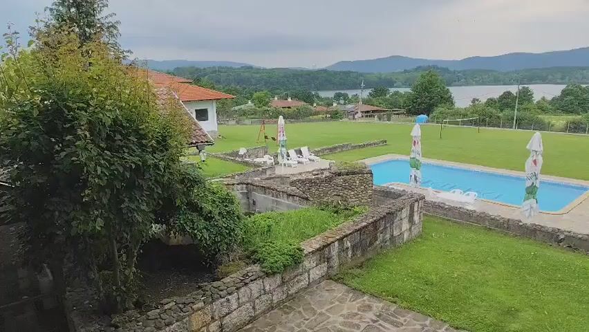 The video displays an expansive outdoor scene featuring a large, meticulously manicured green lawn. In the center of the lawn is a rectangular swimming pool filled with blue water, surrounded by white lounge chairs and four tall, colorful umbrellas, two on each long side. In the foreground, a low stone wall structure with lush green vegetation encloses a small channel of water. To the left, a white building with a terracotta roof is visible. The background reveals a vast body of water, likely a lake or reservoir, with rolling hills and trees beyond it under an overcast sky. A volleyball net or similar recreational structure with a blue ball on top stands on the lawn to the left of the pool. The camera slowly pans from left to right, offering a panoramic view of the property and its scenic surroundings.