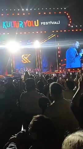 The video shows a large crowd of people at a concert, with a stage in the background. The stage is decorated with lights, screens, and a large "SK" logo. On the screens, there's a close-up shot of a man in a blue suit, presumably the performer. Many people in the crowd are holding up their phones, likely taking photos or videos. The atmosphere seems energetic and celebratory. The video is taken from within the crowd, giving a sense of being present at the event. A sign reads "TÜRKİYE KÜLTÜR YOLU FESTİVALİ".