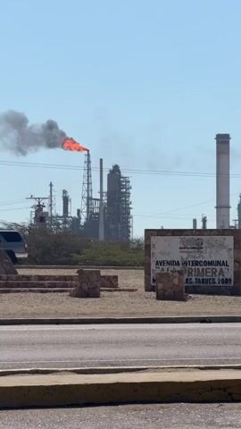 The video shows an industrial complex under a clear, blue sky. A large flare burns atop a tall stack, emitting a plume of dark gray smoke. The complex is comprised of towers, pipes, and various industrial structures. A roadside sign reads "Avenida Intercomunal" in Spanish, with a date of 2009. The area around the sign is dusty and barren, with sparse vegetation. A roadway with a yellow curb is in the foreground.