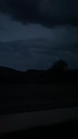 A nighttime video shows a dark landscape with a distant light in the sky. Hills and trees are silhouetted against a cloudy, dark blue sky. A horizontal road or path is visible in the foreground, with a single, small red light appearing briefly on its surface. The light in the sky is small, orange, and appears to be stationary or moving very slowly. The overall mood is dark and atmospheric.