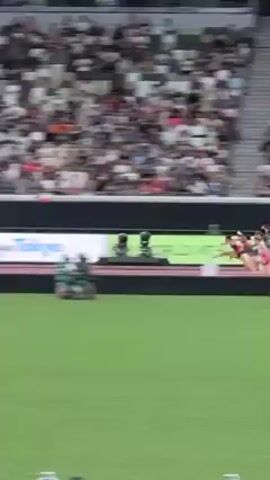 A video captures a running race on a track, with several athletes sprinting forward. The runners, all wearing athletic gear, are spread across the lanes as they race. The crowd is visible in the background, filling the stadium seating. A large electronic display runs along the side of the track, and ground-level filming equipment sits on the grass. The setting appears to be a major athletic competition in an outdoor stadium.
