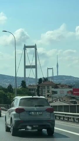 A vertical, handheld shot captures a view from a moving vehicle on a highway. In the foreground, a grey Suzuki Vitara is driving, its license plate visible as 34 HUB 026. Another car, a grey sedan, is in the left lane, further ahead. The road has white lane markings and a metal barrier on the right. In the mid-ground, trees and buildings are visible. Behind them, a large suspension bridge with two towers is prominent. To the right of the bridge, a tall, slender radio tower can be seen. The sky is blue with scattered white clouds. The overall impression is of a scenic drive on a clear day, with urban and infrastructural elements in the background.