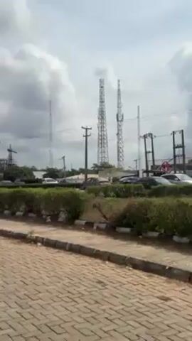 The video shows a paved area in front of buildings with green trees and bushes. There are several tall communication towers visible in the distance.  The camera pans to the right, showing a GTCO sign and a sign indicating "Royal Airport Services." The camera continues to pan, revealing a group of people in the distance walking down the paved area. A large UBA ATM sign is visible, and behind it, a white and gray building is partially visible. In the background, a lush green hill can be seen. The sky is overcast with gray clouds.