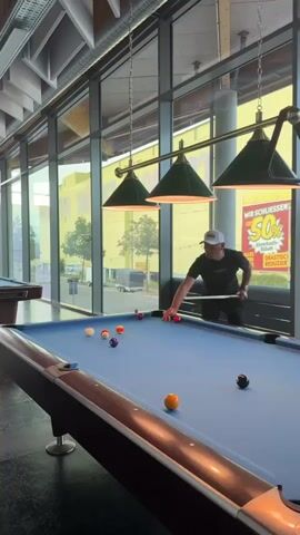 The video shows a pool hall with several pool tables. In the foreground, a blue pool table is visible with several balls on it, including a cue ball. A man in a white hat and dark shirt is standing at the table, holding a pool cue, appearing to play. In the background, another pool table is visible, and through the large windows, a yellow building and trees can be seen. A hand holding a cocktail with straws enters the frame in the foreground, obscuring part of the scene. The lighting suggests it is daytime.