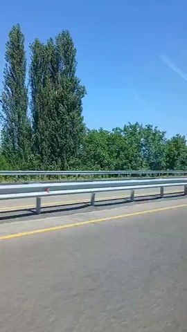 A car is driving on a highway on a clear, sunny day. The camera is positioned inside the car, looking out the window. The highway has a guardrail on the right side, and beyond the guardrail, there are trees and a blue sky. The road is asphalt and has a yellow line marking the lane. A white car is seen passing by on the highway.