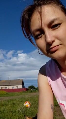 A close-up, handheld video shows a woman in a pink top petting a calico cat outdoors. The woman is in the foreground, her face partially visible, looking at the camera with a neutral expression. The cat is in front of her, facing away from the camera. The woman's hand is stroking the cat's back.

The scene is outdoors, with a grassy area and dirt path. A colorful ball is visible in the background, along with a house and trees under a bright, cloudy sky. The lighting suggests it is daytime, likely late afternoon due to the long shadows. The overall mood is calm and relaxed, with the interaction between the woman and the cat appearing affectionate.

The location is identified as Tashkazgan, Russia. No visible text is present in the video.